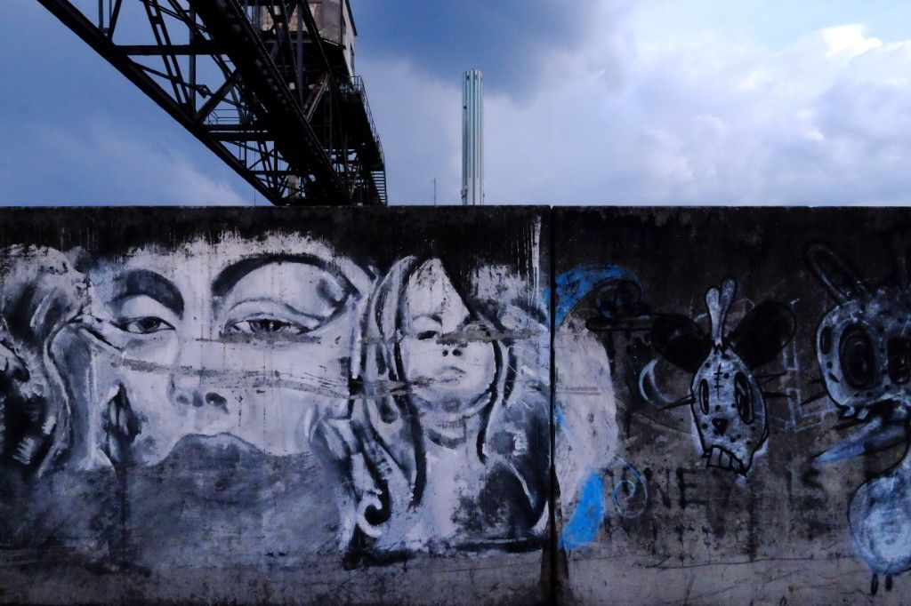 Graffiti of a woman’s face on a concrete wall, with a crane and a chimney behind it