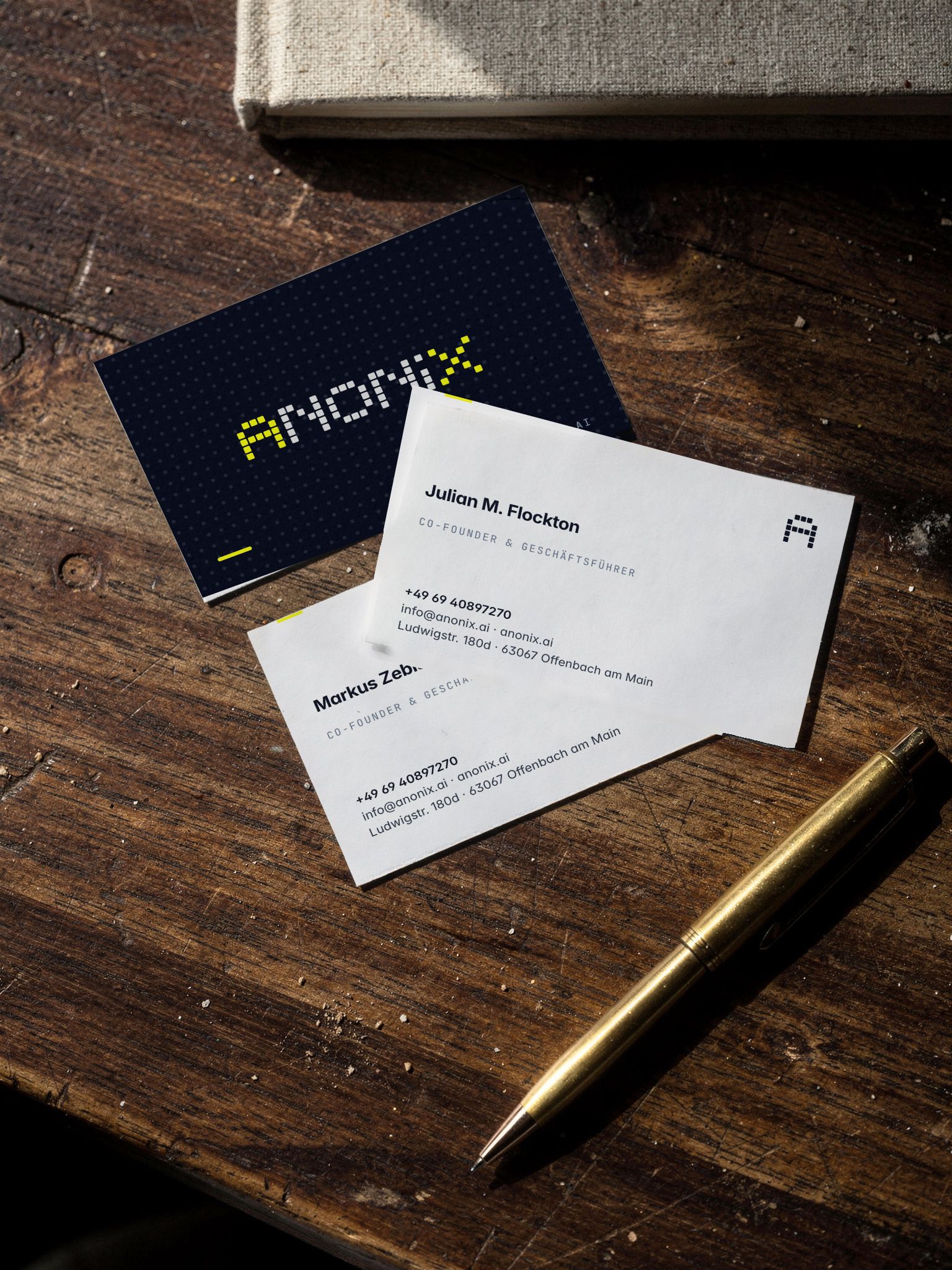 Anonix business cards on a dark wooden table, one dark back with the logo and two white fronts, a gold pen beside them