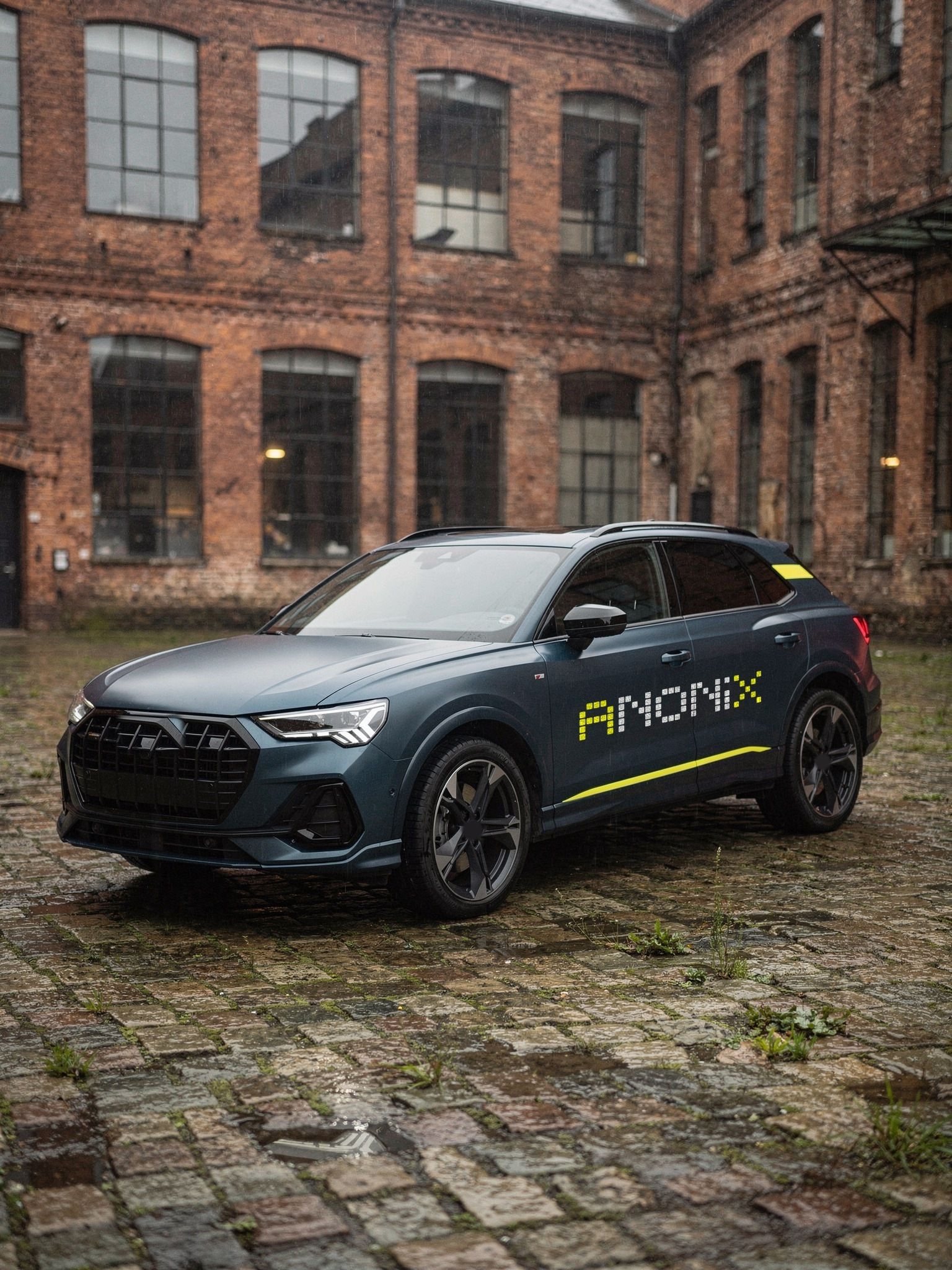 Dark blue SUV in front of an old brick hall on wet cobblestones, the Anonix logo and a yellow line along its side