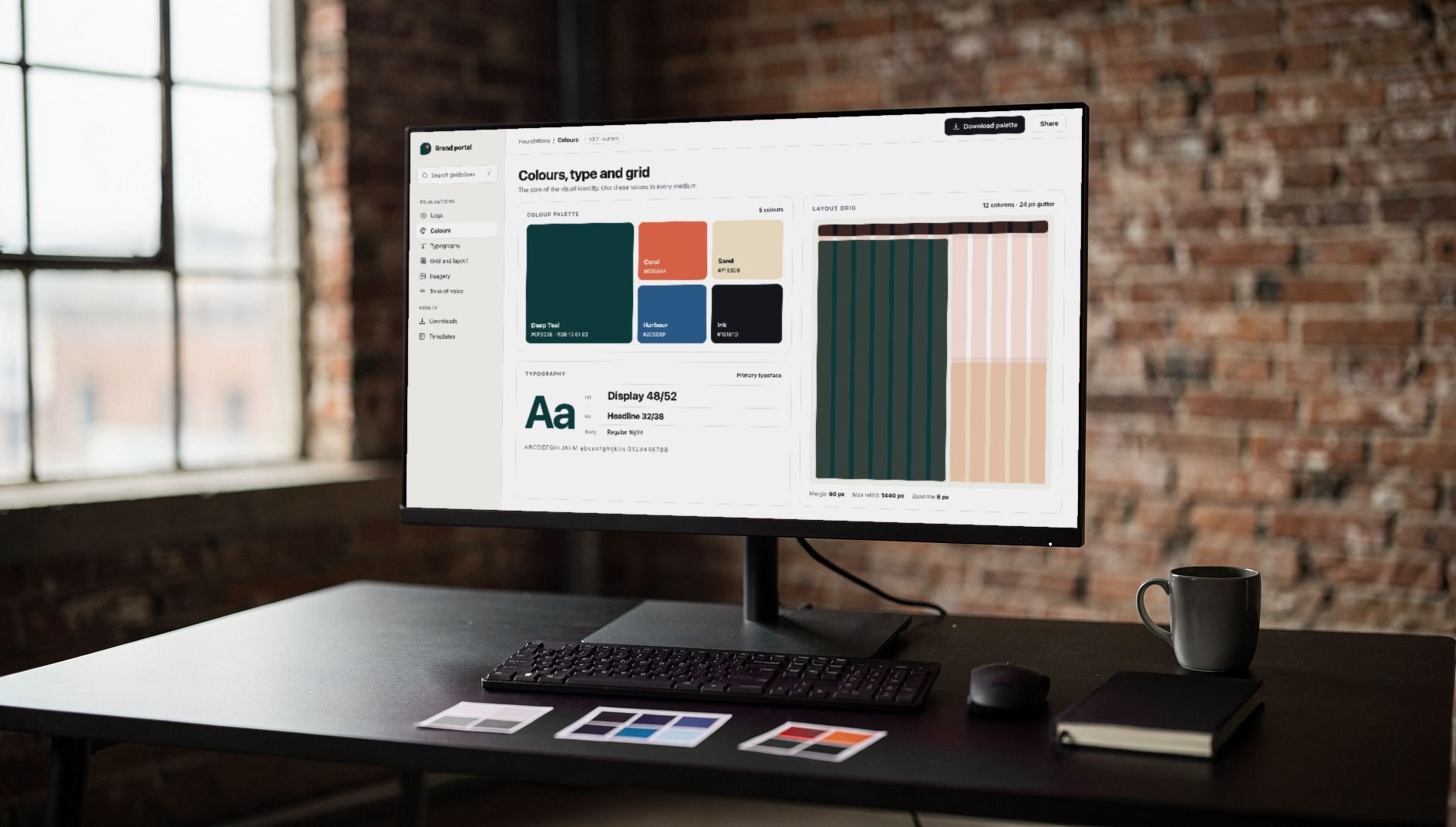 Monitor on a black desk showing a brand portal with the colour palette, type specimens and layout grid, a brick-walled loft behind it