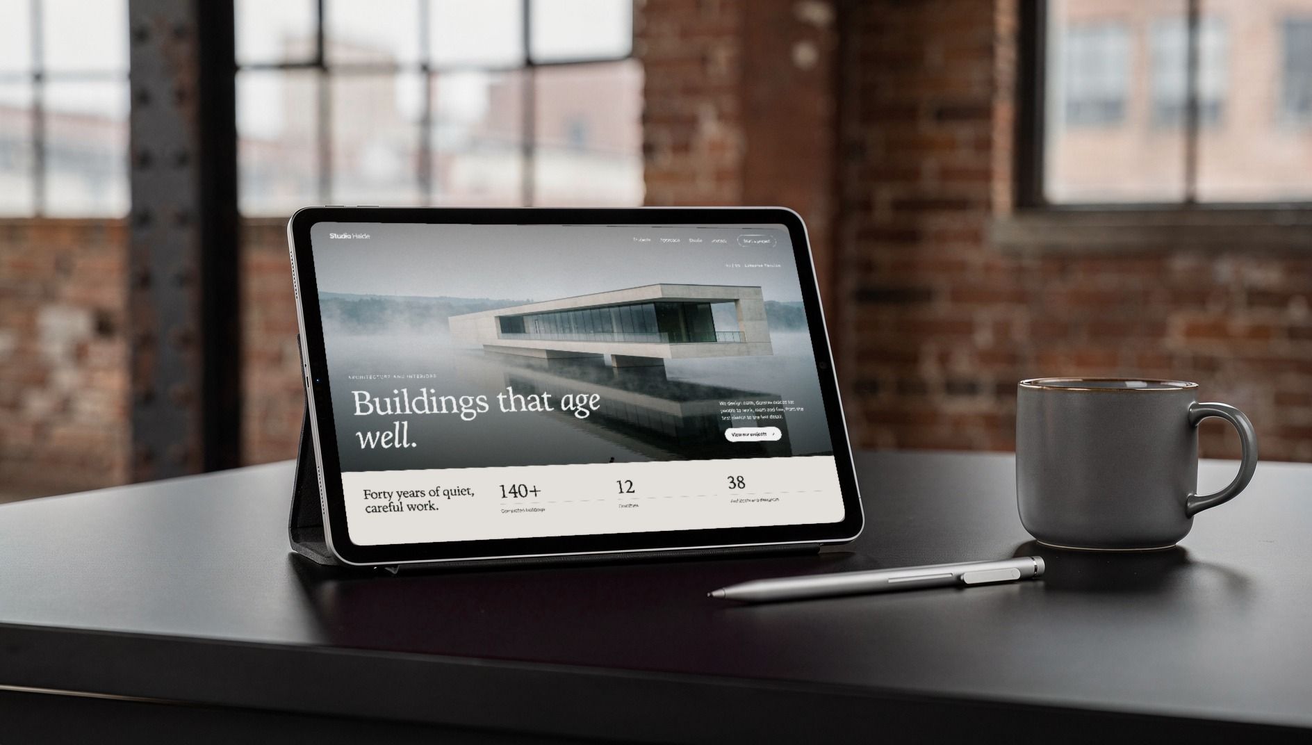 Tablet on a black desk showing the home page of a corporate website with a large architecture photo, a brick-walled factory loft behind it