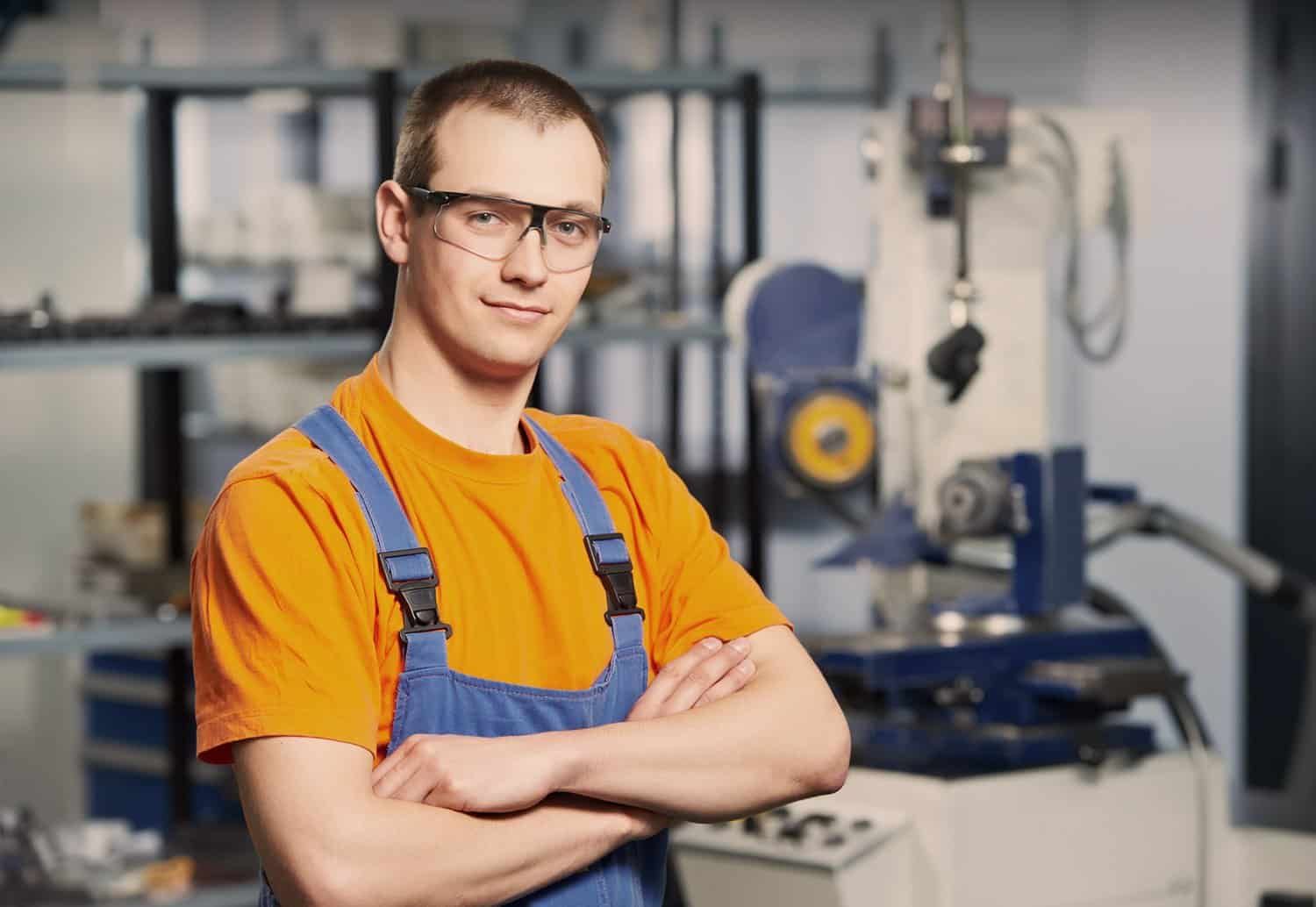 Worker in an orange T-shirt and dungarees wearing safety glasses, arms folded, in front of machines in a workshop