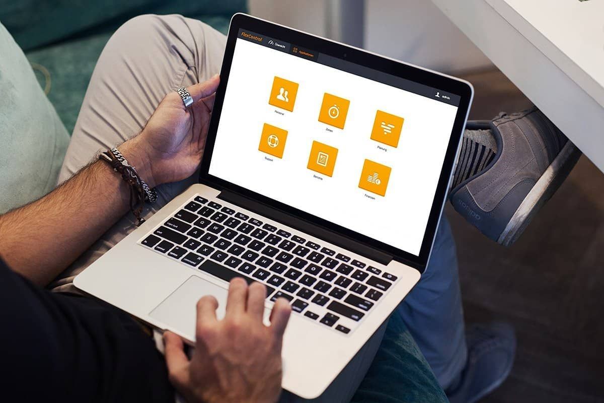 Laptop on someone's lap showing the FlexControl start screen with six applications: Personnel, Times, Planning, Support, Reports and Finance