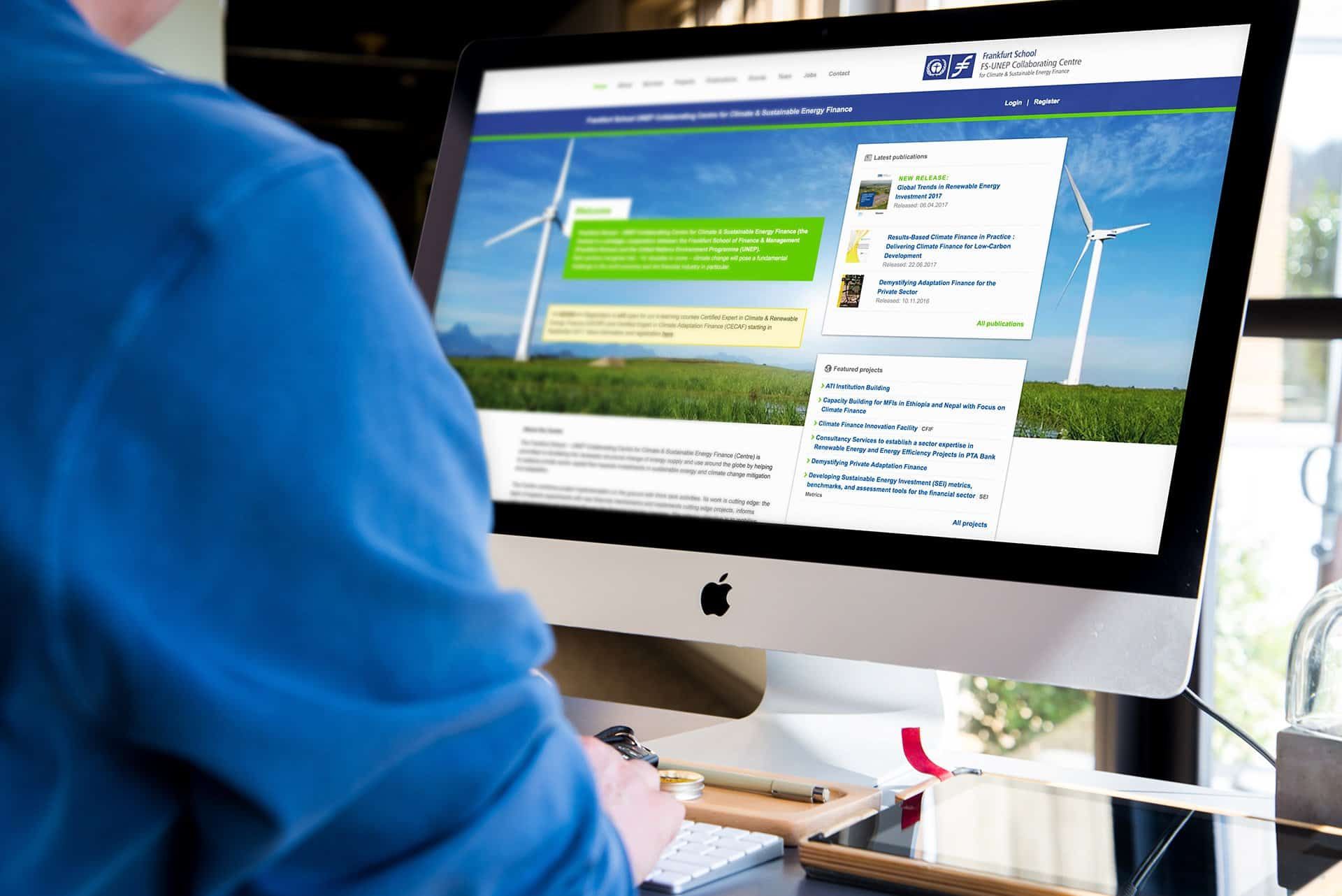 A man in a blue shirt sitting at an iMac showing the UNEP Centre website: wind turbines in the header image, recent publications and projects beside them