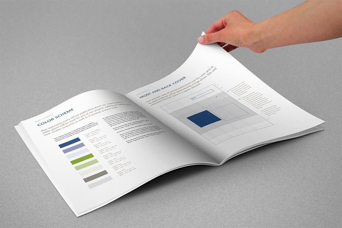 A hand turning the pages of the open design manual: the colour scheme in blues, greens and greys on the left, the grid for front and back covers on the right