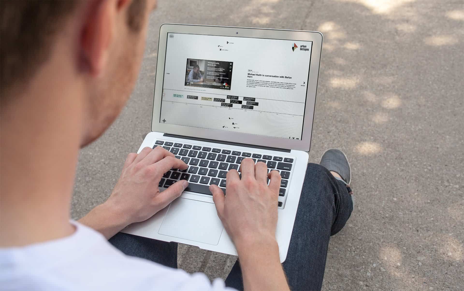 Person with a laptop on their lap, the screen showing the urban-biotopes.net timeline with a video interview