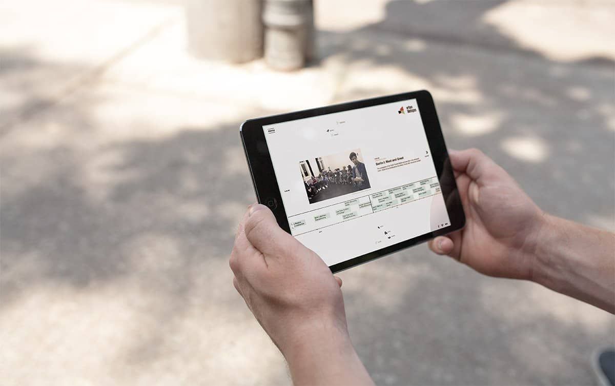Two hands holding a tablet sideways, showing the entry “Berlin I: Meet and Greet” with a photo and the timeline below