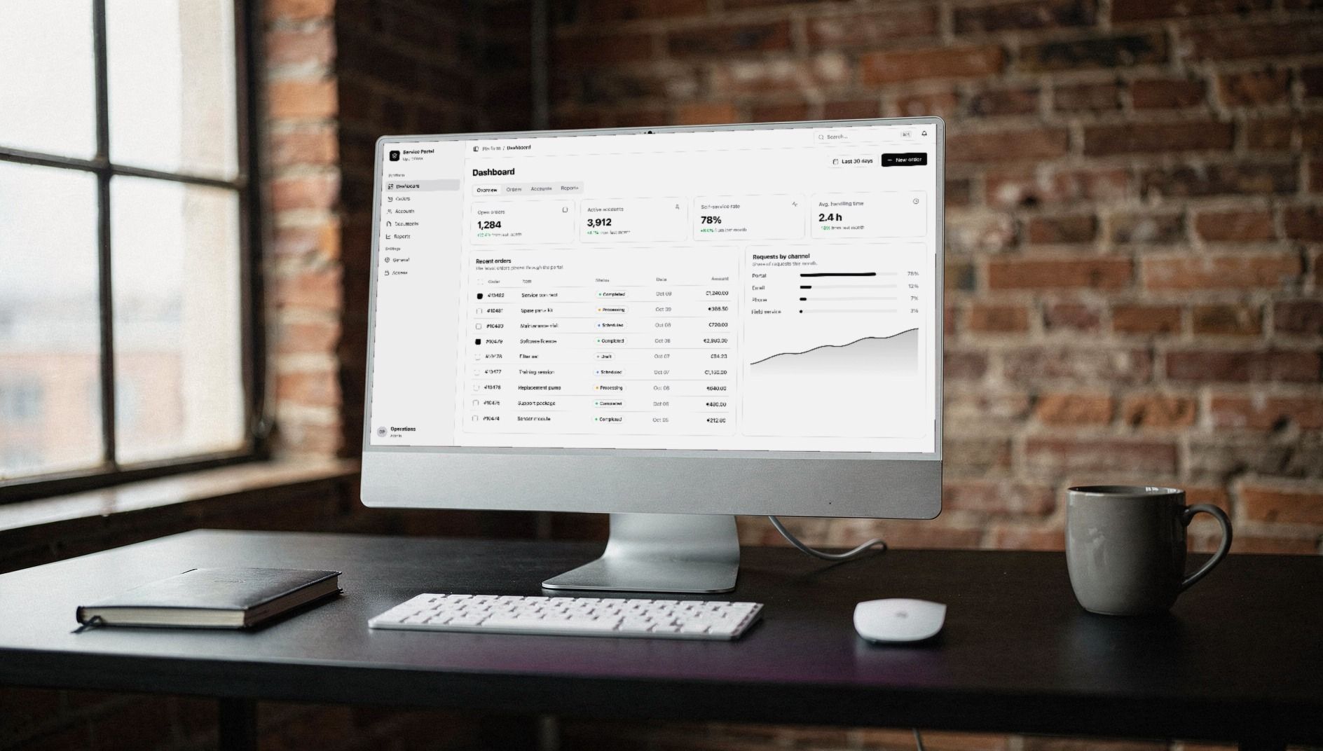 Desktop computer on a black desk showing a portal dashboard with key figures and an orders table, a brick-walled loft behind it
